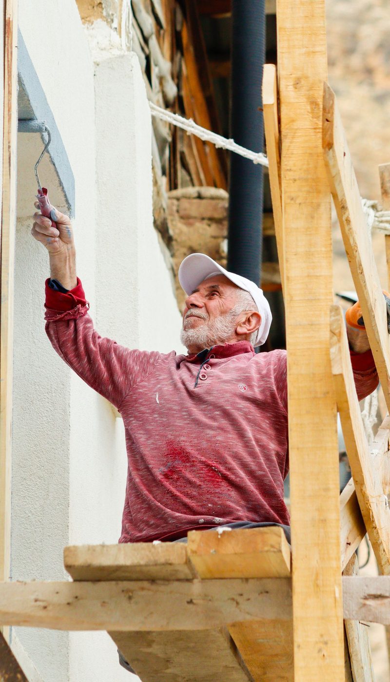 A painter in a red sweater and cap reaching up with a roller to coat the cream render of a house exterior, working from scaffolding in daylight.